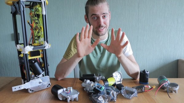 A white male in a green shirt sitting next to a tall rectangular robot made of green and black components with an aluminum frame. In front of him are a variety of components from several windshield wiper motor assemblies. Casings, gearboxes, and the like are strewn across the wooden table.